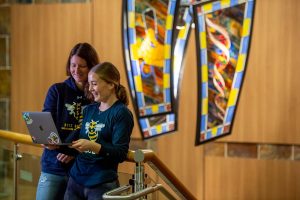 Two young white women wearing navy blue long sleeve RCTC Yellowjacket Soccer shirts smiling at an open laptop