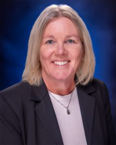 Woman with blond hair wearing a black blazer over a light purple shirt, smiling in front of a blue background