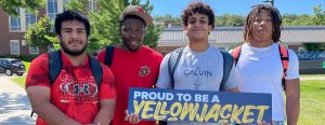 Four RCTC students standing together holding a navy blue sign "Proud to be a Yellowjacket" on a bright sunny late August day