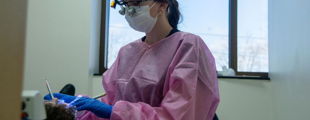 RCTC Dental Hygeine student wearing pink scrub covers, blue latex gloves and white face mask holding dental cleaning tools in a patient's mouth
