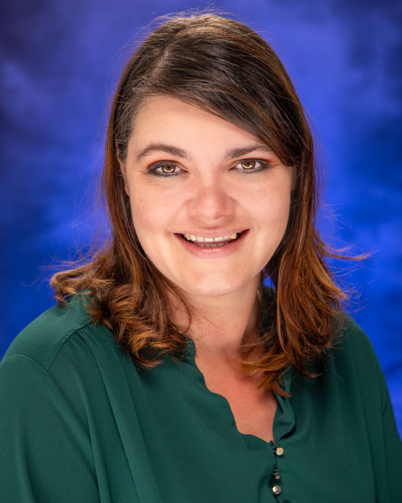 Women with dark brown hair, brown eyes, wearing a dark green blouse against a blue backdrop