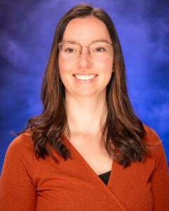 Headshot of Megan Griebenow, white woman with shoulder length brown hair, thin frame eyeglasses, and a burnt orange v-neck shirt sitting in front of a blue background
