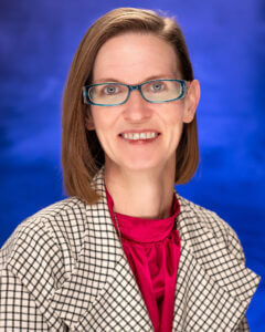 Woman with burnette hair, wearing glasses, a red shirt under a plaid blazer in front of a blue background