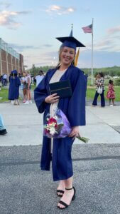 RCTC graduate wearing a navy blue graduate robe and navy blue graduation cap holding a bouquet of flowers.