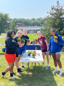 Group of students standing outdoors around a table on the RCTC campus, each holding small potted yellow flowers, with more plants and a watering can on the table.