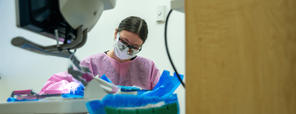 Dentist wearing magnifying glasses and a pink gown focused on a dental procedure in a clinical setting.